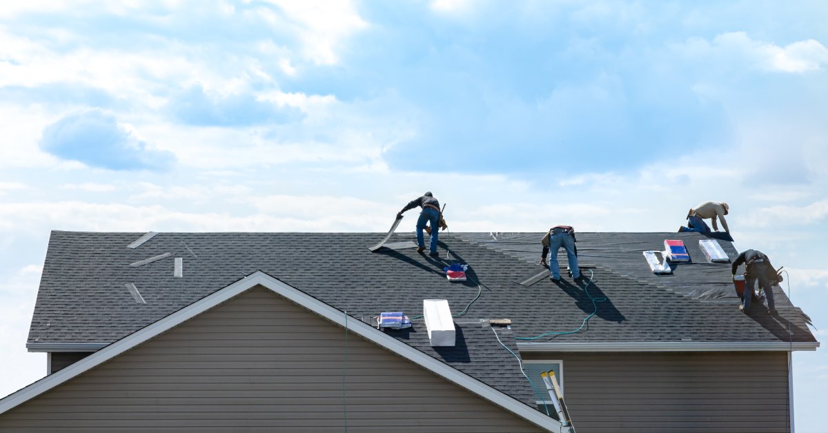 Four different workers stand on a roof and work on installing shingles while the sky behind them is full of clouds.