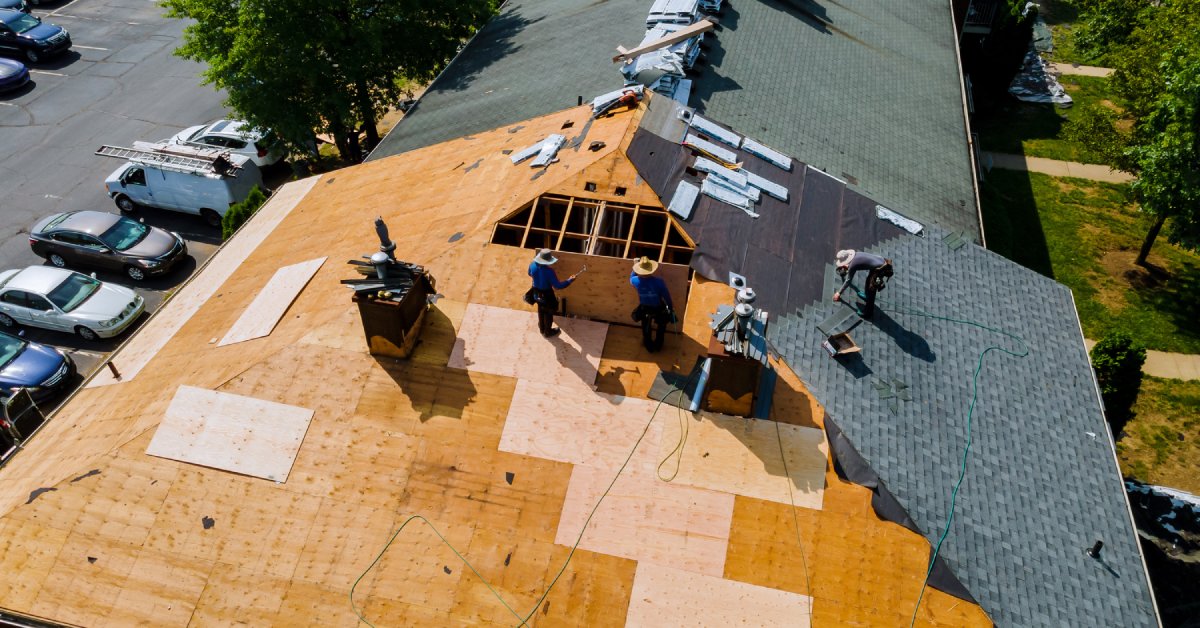 Four different workers wearing hats stand on a large roof while they work on covering it in shingles.