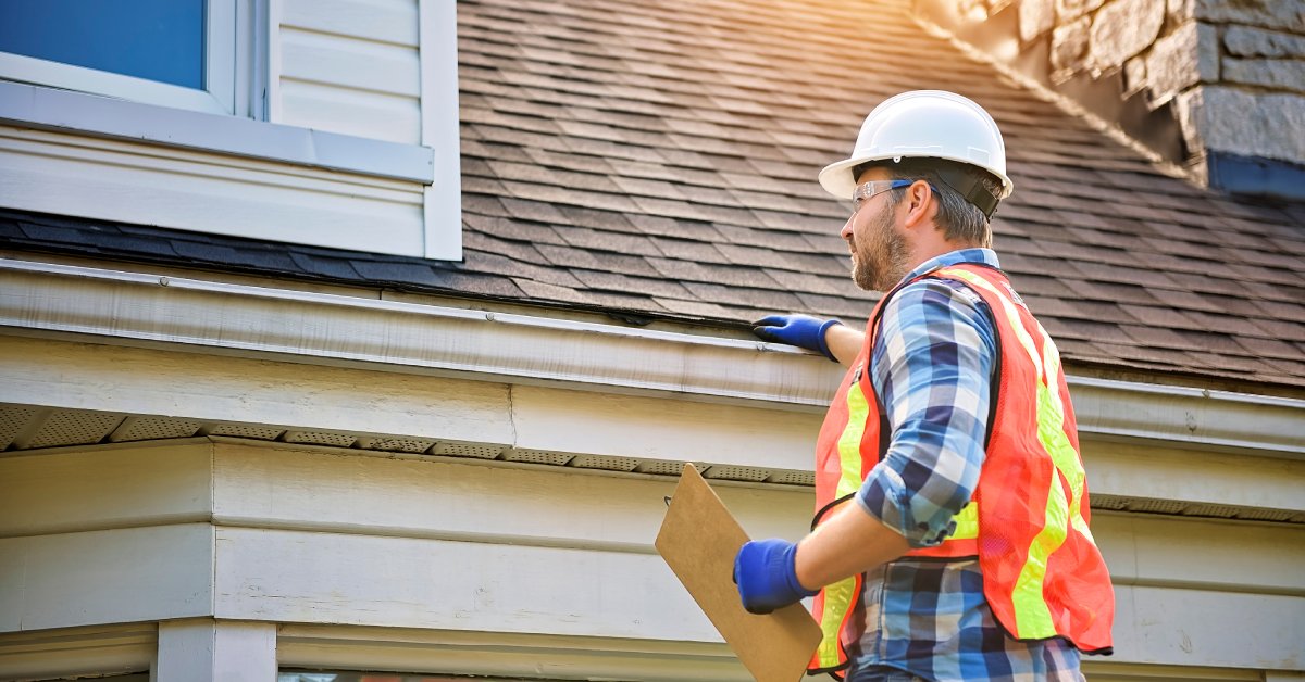 A worker wearing a hard hat and high-visibility vest surveys a roof while they hold a clipboard in one hand.