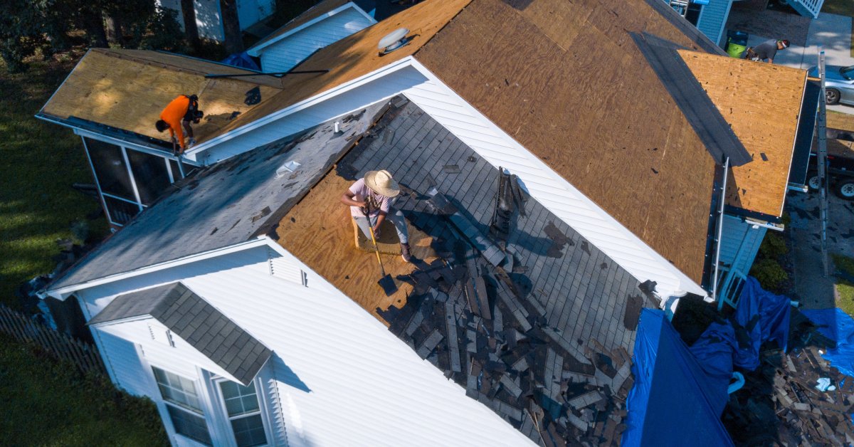 A house in the middle of a roof replacement, with multiple workers scraping off old shingles onto a tarp.