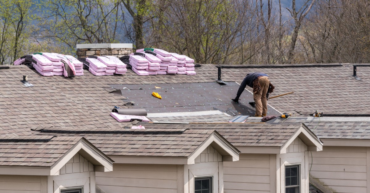 A worker on a large roof using their tools to remove old shingles,. There are multiple stacks of pink bags near the worker.