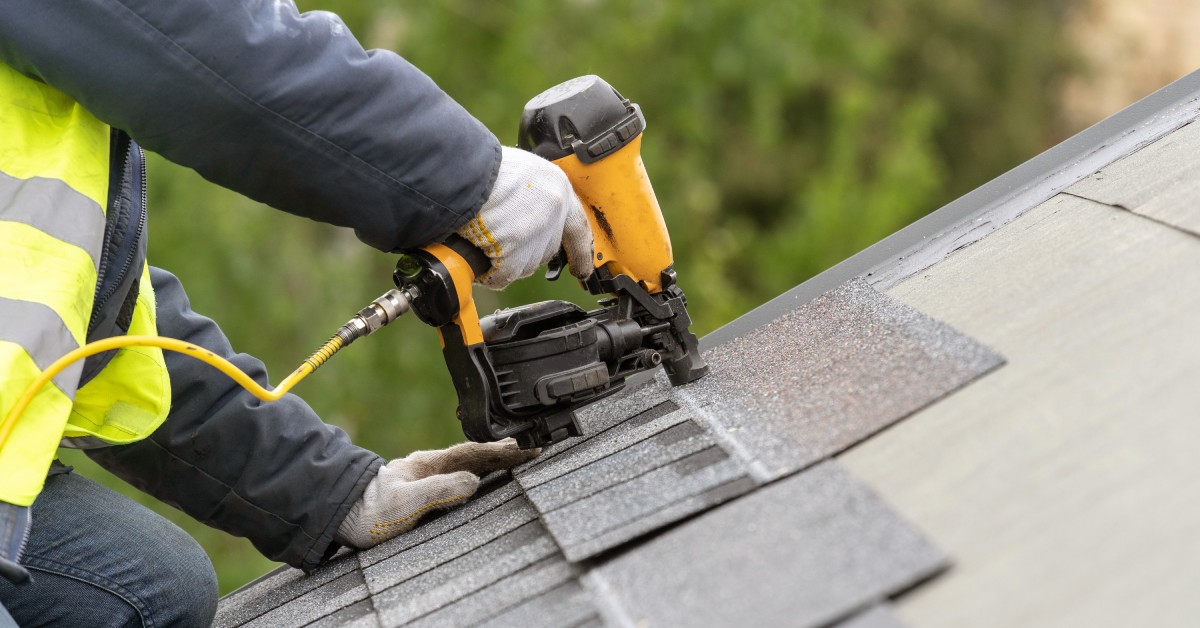 A worker wearing a high-visibility vest is on their knees and using a nail gun to install shingles on a roof.