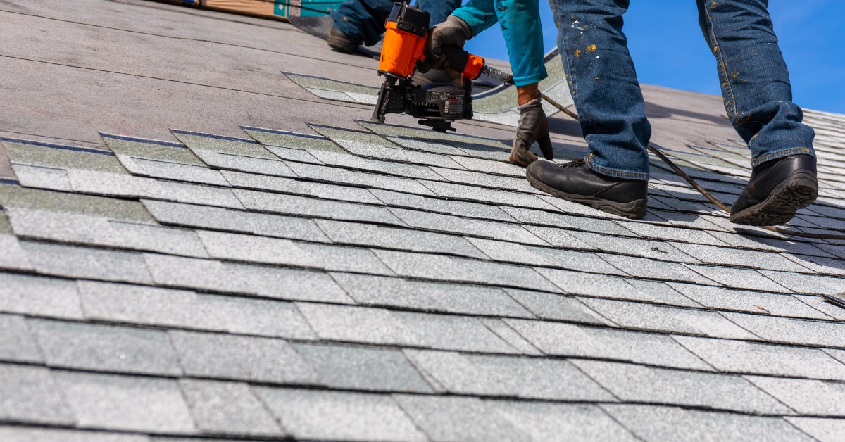 A worker crawling up a roof and and using a nail gun to install shingles. The current shingles are half-finished.
