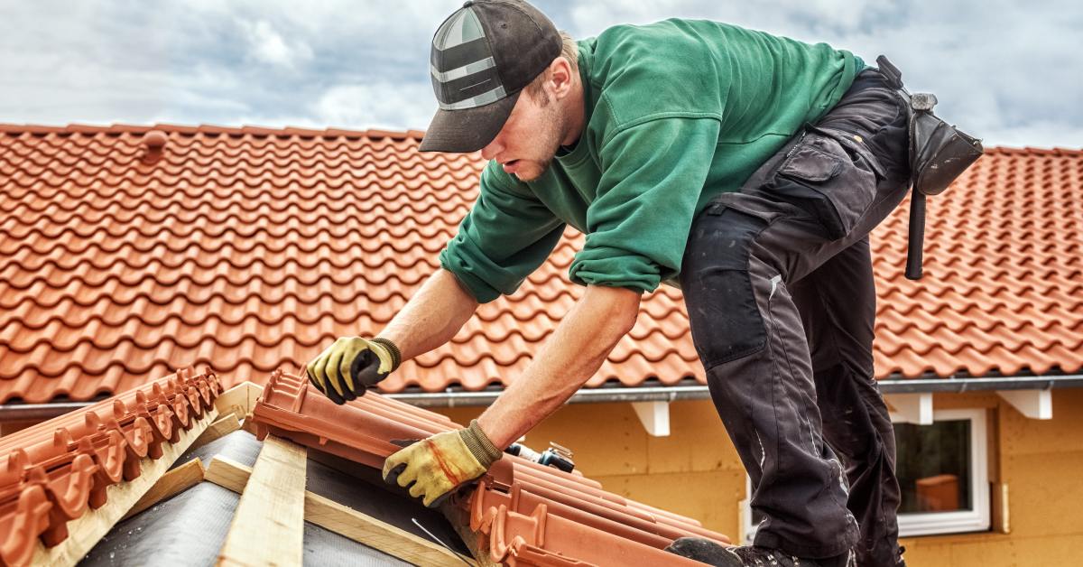 A worker wearing gloves installs new tiles on an orange roof. There is a cloudy gray sky in the background.