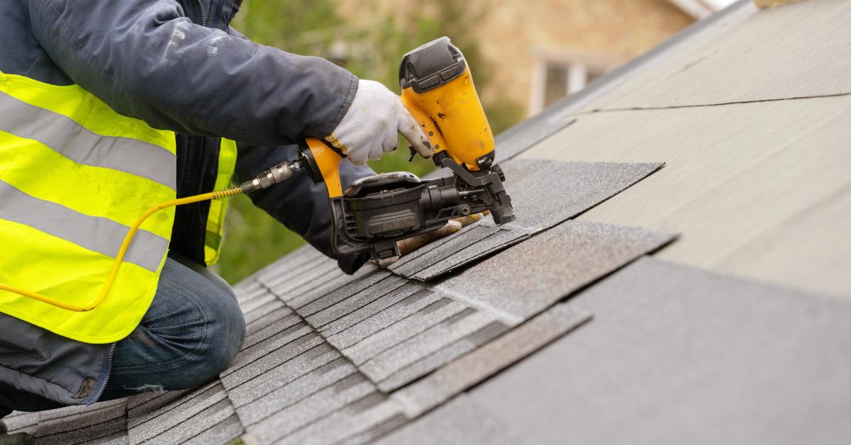 A roofer wearing a protective jacket and gloves while they use a nail gun to install new shingles on a roof.
