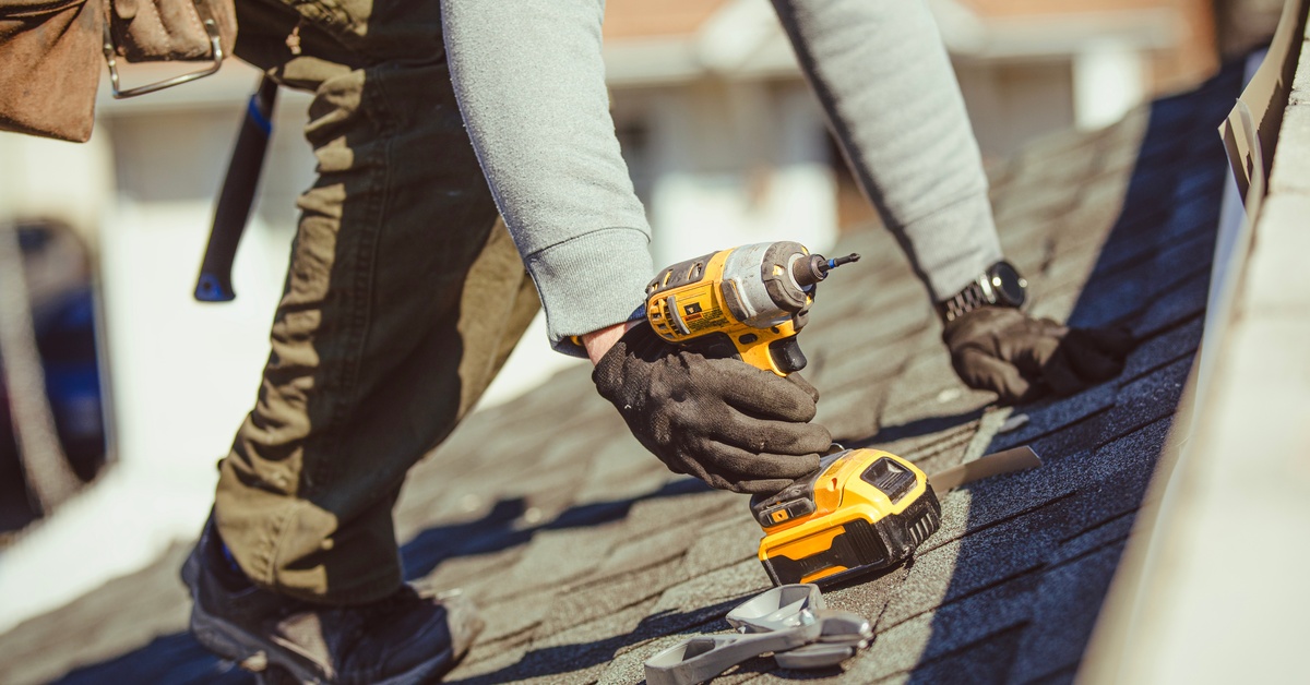 A close-up of a person wearing gloves and holding a power drill on their hands and knees on a shingle roof.