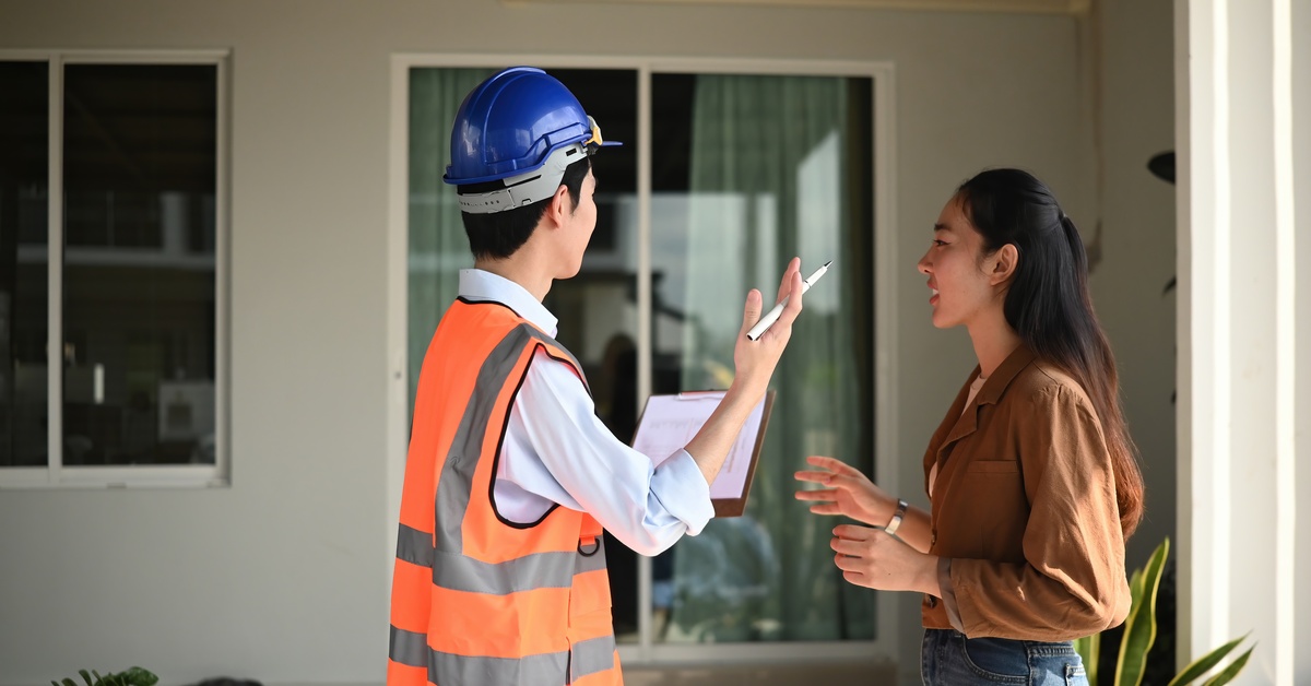A female homeowner stands outside a home with a contractor wearing a safety vest and hard hat, looking at the house.